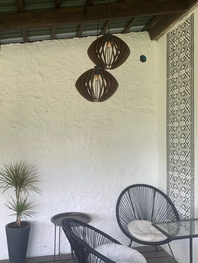 The image displays an unoccupied covered outdoor living space, likely a veranda or patio, located in Yasinia, Ukraine. A prominent textured white wall occupies the left and central background, meeting a corner on the right. This corner features a vertical strip of dark grey geometric patterns on a white background, resembling traditional Ukrainian folk motifs. A small, dark, circular aperture is present on the white wall near the corner. Overhead, a dark wooden beam supports a ceiling structure with visible corrugated roofing material. Two identical, dark brown, oval-shaped pendant lamps, constructed from slatted wood or similar material and containing exposed bulbs, hang from the ceiling. The floor is composed of dark wooden planks. Furnishings include two black, round, open-weave "Acapulco-style" chairs, each fitted with a white circular cushion. A dark, round, elevated side table with a tray-like top is positioned between the chairs. A portion of a round glass-top table with a dark frame is visible on the far right. A tall, slender Dracaena plant in a dark grey conical planter stands on the floor on the left side of the scene. No individuals or actions are depicted.