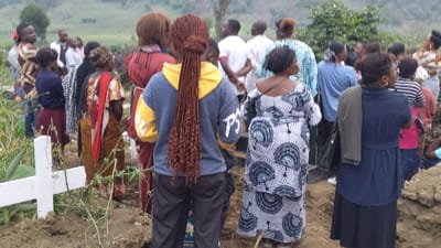A gathering of people, mostly women, stand around what appears to be a gravesite marked by a white cross. They are dressed in various attire, some in traditional patterns and others in contemporary clothing. The focus is on their backs as they face the grave, suggesting a somber occasion, possibly a funeral or memorial service. The setting is outdoors with green vegetation visible in the background.