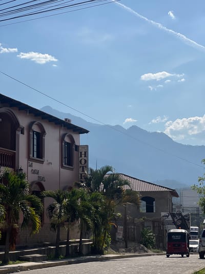 The image shows a street scene in Gracias, Honduras, with a building on the left side labeled "La Casa Real Hotel". Palm trees are growing along the sidewalk in front of the hotel. In the background, a large mountain is visible. On the right side of the image, a row of parked vehicles and a red and black tuk-tuk are visible along the street. The sky is blue with scattered white clouds and contrails. Overhead power lines run across the sky.