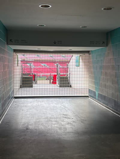 This media file captures an interior view from a stadium concourse or hallway, looking out onto the seating area and pitch. The perspective is from a relatively dim corridor that opens into the brighter stadium bowl through a black metal grated barrier.
On the left side of the corridor, a tall, smooth grey wall is visible, featuring several circular recessed lights. This wall displays multiple signs indicating seating directions. The upper, more distant sign reads "ROWS SEATS 31-44 95-110" with an arrow pointing right, towards the stadium. Closer to the viewer, another sign reads "ROWS 02-30" with an arrow pointing left. A third sign, partially visible further left, indicates "ROWS SEATS 31-44 111-124" with an arrow pointing left. The corridor floor is composed of dark grey and greenish-blue rectangular tiles. The ceiling in the upper background (left side) features light blue and grey rectangular tiles. The right side of the corridor is a textured, dark grey concrete wall.
Through the metal grating, the vibrant interior of a stadium is revealed. Numerous rows of bright red seats are arranged in tiers, descending towards a green sports pitch, a section of which is visible at the lowest level. Support structures for the seating and a small, greenish-tinted window are also visible in the distance. The stadium is empty, with no people present. The lighting indicates it is daytime. Based on the provided context, this scene is set within Wembley Stadium in the United Kingdom.