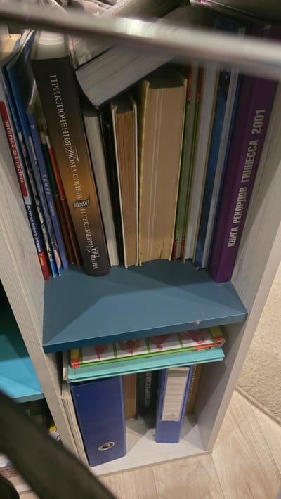The image shows a bookshelf filled with various books and binders. On the top shelf, there are several books with their spines facing outwards. One book has Russian text that appears to read "Приключения Тома Сойера" (The Adventures of Tom Sawyer). Another book has the title "КНИГА РЕКОРДОВ ГИННЕССА 2001" (Guinness World Records 2001). Below this shelf, there is another shelf with what appears to be binders and more books. One blue binder is prominently visible. The bookshelf itself is made of light-colored wood with a blue shelf insert. The surrounding environment is not clearly visible but appears to be an indoor setting with a carpeted floor. There are no people or animals depicted in the image. The scene is likely a domestic or office setting, the exact location in Ukhta, Russia, is unknown.