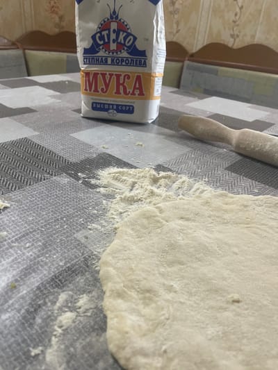 The image depicts a close-up, angled view of a table surface covered with a checkered tablecloth. On the left side, a white and blue bag of flour labeled "СТ. КО МУКА" is partially visible. The word "МУКА" is prominently displayed in large yellow lettering. Below this, text in smaller font reads "СТЕПНАЯ КОРОЛЕВА" and "ПШЕНИЧНАЯ ХЛЕБОПЕКАРНАЯ ВЫСШИЙ СОРТ".
To the right of the flour bag, a wooden rolling pin dusted with flour lies on the tablecloth. Adjacent to the rolling pin, a section of light-colored dough is spread out, also dusted with flour. Scattered flour is visible on the tablecloth around the dough and rolling pin, indicating recent activity.
The background shows a wall with a floral wallpaper pattern and a wooden shelf or furniture. The lighting suggests an indoor setting, possibly during the daytime. The overall scene implies food preparation, specifically baking, is in progress. The location context provided is Линёвка, Russia. There are no people visible in the image.