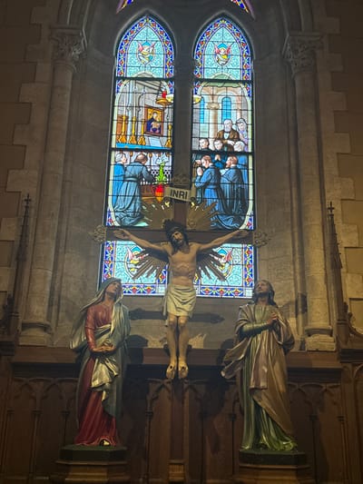 The image shows a depiction of the crucifixion inside a church. Jesus Christ is mounted on a cross with the inscription "INRI" visible. Flanking the cross are statues of religious figures, one dressed in green and gold robes, the other in red and grey robes. Behind the cross and statues is a stained-glass window featuring scenes of religious figures. The scene is illuminated by soft, diffused lighting.