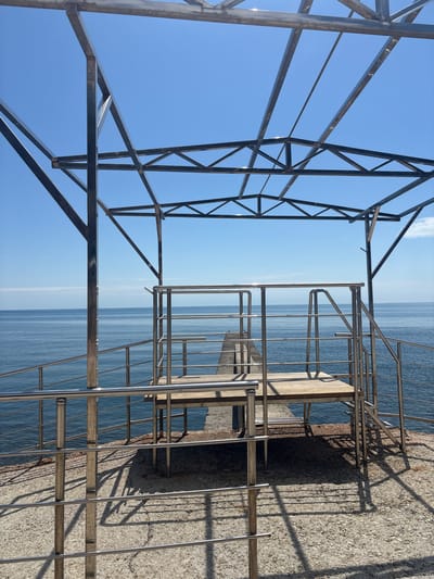 The image shows a pier structure under construction in Semidvorye, Ukraine. Metal scaffolding frames the left side of the image, reaching towards a blue sky with minimal clouds. To the right, a finished section of the pier features metal railings and a short staircase leading down to the sea. The sea, a pale blue, stretches to the horizon. A concrete pier surface occupies the right edge of the frame, casting shadows from the railings onto its textured surface. No people are visible.