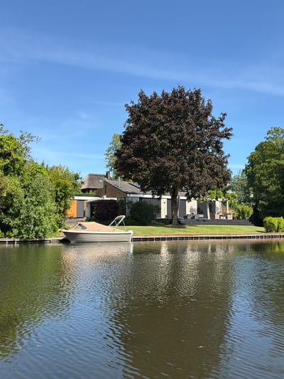 A sunlit scene shows a narrow waterway in Leerdam, Netherlands. The waterway is flanked by a timber retaining wall, and the water’s surface has gentle ripples. Alongside the water, on the left side of the frame, are dense green bushes and a dark-leaved tree. Behind the bushes and tree, a structure with a thatched roof is visible. A light-colored boat, covered in a tarp, is docked next to the retaining wall. On the other side of the thatched building, a modern structure is partially obscured by a large tree with reddish-brown leaves. In the background, a row of gravestones is visible across a manicured lawn. The sky is clear blue with faint clouds.