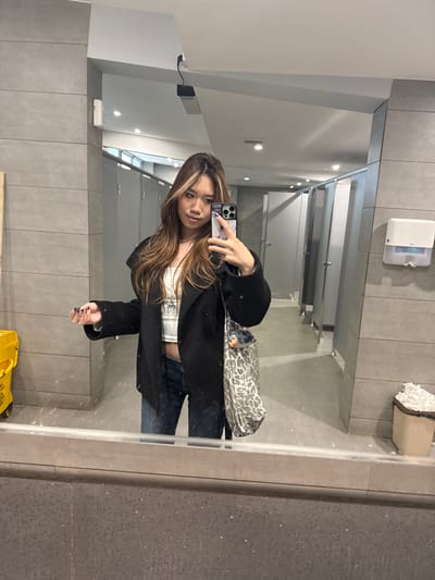 A person is taking a mirror selfie in what appears to be a public restroom. The person has long, brown hair with blonde highlights, and is wearing a black coat, a white patterned top, and blue jeans. They are holding a smartphone in their left hand to take the picture. A patterned bag is slung over their shoulder.

The background of the photo shows a public restroom with gray tiled walls and several bathroom stalls. A paper towel dispenser and a waste bin are visible on the wall above. The floor has a yellow safety sign. The lighting in the restroom appears to be fluorescent.

There is no indication that this photograph was taken in Guazhou County, China.