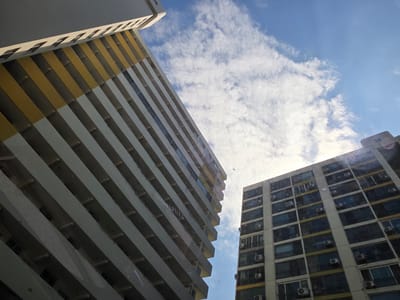 The image captures a view of apartment buildings in Eumseong-eup, South Korea, from a low angle looking upwards towards the sky. Two prominent apartment buildings dominate the frame. The building on the left is taller and features a distinctive yellow and white facade with numerous balconies. The building on the right is also an apartment complex, characterized by its repetitive window patterns and external air conditioning units. The sky above is a bright blue with scattered white clouds, illuminated by sunlight that creates lens flares in the upper right portion of the image. No people are visible in the scene. The buildings appear to be residential structures within an urban or semi-urban setting.