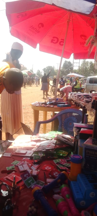 This image depicts an outdoor market scene in Chemuche ward, Kenya, under a bright, sunny sky.
In the immediate foreground, a red market umbrella bearing reversed "Airtel" and "Airtel Money" branding provides shade. Below the umbrella, a person with dark skin, wearing a white kufi or cap, a yellow vest over a white garment, and a long, light-colored skirt, is bent forward, looking down at a display table. A metallic cross pendant hangs from their neck.
The foreground table, covered with a red surface, holds an assortment of small items for sale. These include numerous packets of mobile phone screen wipes, red and white SIM card packs (some with a "G" logo), multiple colorful plastic flashlights (blue, pink, orange, yellow), USB cables, padlocks with keys, and various small electronic accessories. To the right, a dark bottle and a box labeled "PREMIER UNIVERSAL W.K." are visible.
In the mid-ground, a round, light-colored plastic table is visible, beside which a blue plastic chair is positioned. Another individual in a light-colored top is bent over near this table, appearing to tend to items on the ground.
The background reveals a bustling, unpaved area of reddish-brown earth, characteristic of a roadside market. Multiple individuals are present; one person in a dark top and light skirt has their right arm raised. Several motorcycles (boda-bodas) are visible, along with a white van or car on the far right. One person on a motorcycle wears a yellow reflective vest. Distant buildings with brown roofs, trees, utility poles, and power lines complete the background environment. Other market stalls with various colored umbrellas can be seen further in the distance.