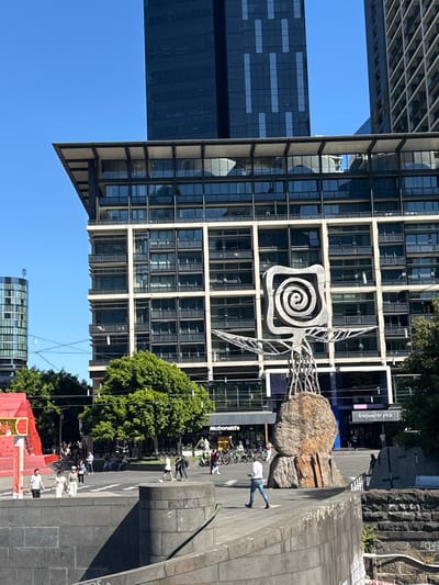 An office building with a spiral symbol on its face dominates the view. Next to it is an art installation featuring a large stone. A street scene unfolds below, with trees, a McDonald's restaurant, people, and bicycles. A clear blue sky is overhead. In the background, another office tower is visible.