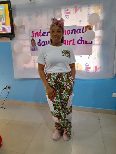 The image depicts an indoor scene in the city of Ajah, Nigeria, featuring a single adult female standing in front of a decorated wall.
The woman, with medium-brown skin, stands centrally, facing the camera with a smile. She is wearing a plain white short-sleeved t-shirt that features a small, rectangular pocket on the left chest, patterned with green, brown, and white floral or leafy motifs. Her lower attire consists of loose-fitting trousers patterned with similar green, brown, and white floral/leafy designs, interspersed with solid white fabric panels on the thighs and shins. On her head, she wears a multi-colored fabric headwrap, primarily pink, light blue, and white, tied into a prominent bow-like knot at the crown. Her footwear consists of light pink, closed-toe slip-on shoes. Her left hand is resting in her trousers pocket, while her right arm is slightly obscured.
The background features a wall painted light blue on the lower section and white/off-white on the upper section. A large white banner or sheet is affixed to the upper wall, displaying the words "International day of girl child" in dark pink lettering. A small portrait of a young girl is attached to the banner on the left, and a small image of a doll or cartoon character is attached on the right. Several partially deflated white balloons are taped or attached to the banner and the adjacent wall. Behind the translucent banner, various logos, patterns, and reversed text are faintly visible on the wall. A dark-framed flat-screen television is mounted on the upper left wall, displaying a colorful image with yellow and blue tones. Below the television, a black electrical cable is plugged into a white wall socket on the light blue wall. Another white electrical socket is visible further to the right. The floor is covered with large, light-colored square tiles.
