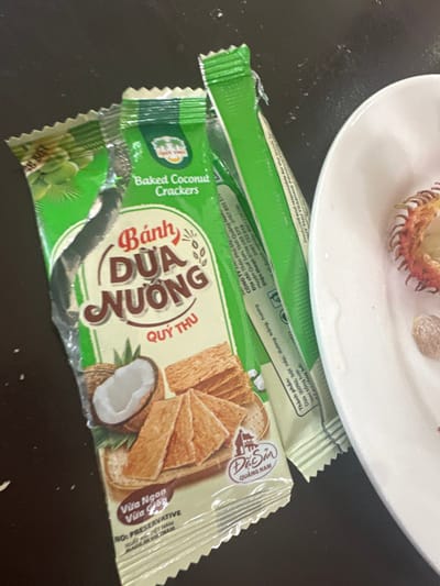 The image shows two packets of "Bánh Dừa Nướng Quý Thu" baked coconut crackers from Quang Nam, Vietnam. The wrappers are green and silver, featuring images of coconut crackers and a half-cut coconut. The text mentions the product is preservative-free, contains 60% coconut, sticky rice flour, sugar, and vanilla flavoring.  Company details and contact numbers are also printed on the wrappers. One of the packets is partially torn open. A white plate with some food scraps sits on top of the packets.