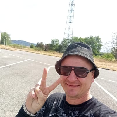 The image is a close-up selfie of a man wearing a dark bucket hat and sunglasses. He is smiling and holding up two fingers in a peace sign. He is wearing a black t-shirt with a dark graphic design. A watch is visible on his left wrist. The background appears to be an outdoor setting, possibly a parking lot, with white lines painted on the asphalt. Beyond the parking lot, there is dry grass, trees, and a hill in the distance under a bright, overcast sky. A tall, metallic tower structure is visible to the right of the man's head, extending upwards towards the sky. The lighting suggests it is daytime. The image was taken in Pamiers, France. No visible text is present in the image.