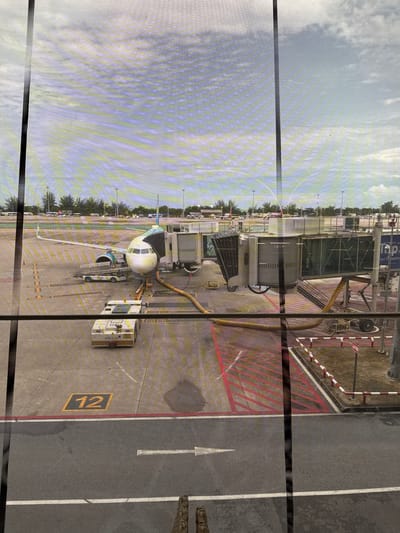 The image displays an airport apron scene captured from an elevated interior perspective, likely within a terminal building at Phuket International Airport (HKT) in ตำบล ไม้ขาว, Thailand. The view is partially obscured by window mullions and a subtle grid pattern, possibly from a screen or reflective glass.
A narrow-body passenger aircraft, featuring a white fuselage with teal or blue accents on the tail and engine nacelles, is parked at gate 12. The aircraft's nose points towards the viewer's left. A flexible, grey jet bridge is connected to the forward left passenger door. Orange and yellow hoses are attached beneath the aircraft, indicating active fueling operations.
Various pieces of ground support equipment are positioned around the aircraft:
* A white and yellow pushback tug, marked "AOTGA AT-014," is situated in front of the aircraft's nose.
* A baggage loader/belt loader, colored yellow and white, is positioned near the aircraft's cargo hold area.
* Other service vehicles are partially visible near the jet bridge and in the background.
The concrete apron surface prominently displays "12" in yellow within a black square, marking the gate number. Red parallel lines are painted on the apron near the jet bridge, indicating safety zones. A grey asphalt road with white markings runs across the foreground. Dark, wet patches are visible on the apron surface.
In the mid-ground, a line of green trees separates the apron from a distant area containing parked cars and vans, suggesting an airport access road or parking lot. Further in the background, other airport structures and potentially another aircraft are visible. The sky above is partly cloudy with patches of blue and white. The very bottom of the image shows a partial reflection of shoes, reinforcing the indoor viewpoint. No identifiable people are clearly visible in the scene.