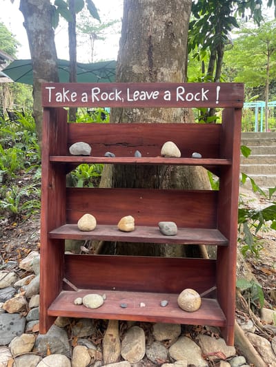 The image shows a wooden shelf with the words "Take a Rock, Leave a Rock!" painted on it, positioned outdoors in a natural setting. The shelf has four compartments, each containing several rocks of varying sizes and shapes. The shelf is placed in front of a tree, with vegetation and rocks visible in the background. The scene appears to be in a park or natural area with a sign requesting visitors to either take a rock from the shelf or leave one behind. It is a "take one leave one" shelf for rocks. The location is inferred to be Ninh Hoa, Vietnam.
