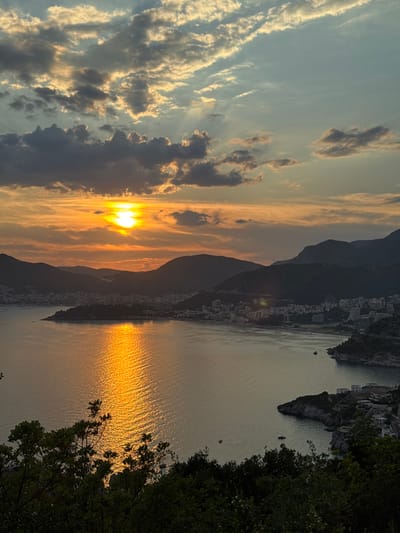 An aerial, long shot captures a sunset over a bay in Čelobrdo, Montenegro. The sun, a bright orange orb, is partially obscured by dark, textured clouds on the left side of the frame. Rays of sunlight pierce through the clouds, casting a golden glow across the sky. The sea below reflects the vibrant sunset, creating a shimmering, rippled path of light. In the mid-ground, dark, silhouetted mountains form a rugged coastline. A city with numerous buildings, ranging from low-rise structures to multi-story hotels, is nestled along the waterfront and up the hillsides. The foreground is dominated by the dark silhouettes of lush green trees and foliage. The overall scene conveys a serene and picturesque coastal landscape at dusk.