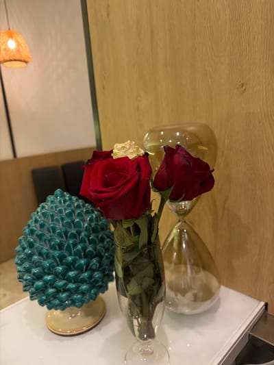A still life arrangement sits on a white surface. In the foreground, a clear glass vase holds three red roses, one with a dried yellow petal. To the left of the roses is a turquoise ceramic object shaped like a pinecone, with a light-colored base. Behind the roses, a large, amber-colored glass hourglass stands on its side. The background consists of a light-colored wall on the left and a warm-toned, wood-paneled wall on the right. A woven lampshade is visible in the upper left corner. The overall scene appears to be indoors, possibly in Otranto, Italy, given the decorative ceramic style.
