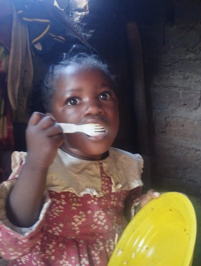 A young Black child is eating from a yellow bowl with a white plastic fork. She wears a dress with a floral pattern in red and white, featuring a ruffled collar. The background includes what appears to be the interior of a rustic structure, with rough walls and assorted objects partially visible. The child is looking upwards with wide eyes.