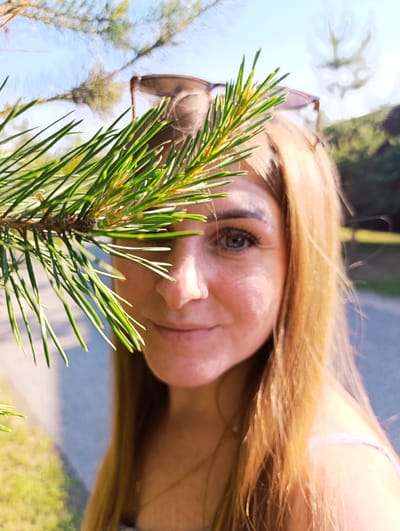 A woman with blonde hair and blue eyes is partially obscured by a pine branch in the foreground. She is wearing sunglasses on her head. The background is a blurred outdoor scene with trees and a path. The image appears to be taken on a sunny day. The location is not discernible from the image itself.
