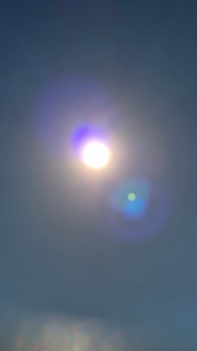 This media file depicts a bright, glowing object, likely the sun, in a hazy or overcast sky. The sun is rendered as a large, luminous sphere with a strong purple and yellow halo. To the upper right of the sun, a smaller, fainter blue and green circular lens flare is visible, suggesting the light source is strong and possibly being captured at an angle through a lens. The sky itself is a muted, gradient blue-grey, with some subtle variations in tone. There are very faint, almost imperceptible speckles throughout the image, which could be dust on the lens or atmospheric particles. The overall impression is one of bright, diffuse light in a cloudy or hazy atmosphere. The setting appears to be outdoors, with no discernible landscape features or human presence. The time of day is not explicitly indicated but the bright light suggests daytime. The emotional tone is neutral, with the focus being on the light phenomenon. There is no visible text in the image.