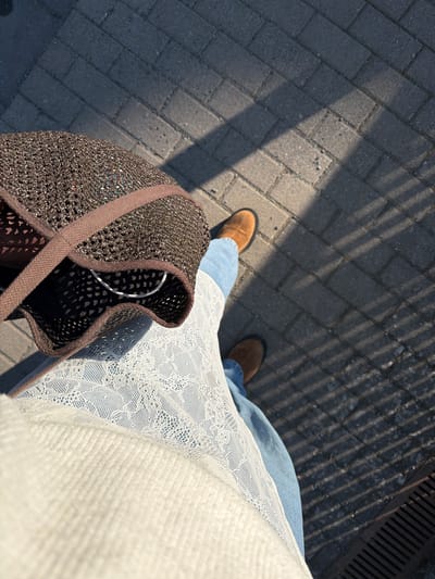 The image, taken from a high-angle perspective looking down, captures a person standing or walking outdoors on a sunny day in Odernheim am Glan, Germany.

The main subject is the lower half of a person, dressed in blue jeans, which are partially covered by a white garment. This white garment appears to be layered, with a delicate white lace fabric visible over what looks like a plainer white knitted or woven material. The person is wearing light brown, possibly suede, ankle boots. Their feet are positioned slightly apart, suggesting either a pause during walking or a pose for the photo.

The person is holding a dark brown, intricately woven or crocheted bag with a matching brown fabric strap, visible in the foreground on the left. Inside the bag, a white hanger can be faintly seen, suggesting it might contain clothes or other items.

The ground is paved with grey rectangular interlocking blocks or pavers, showing some wear and tear. Strong sunlight casts distinct, elongated shadows across the pavement, indicating a fence or railing above and to the right, creating a striped pattern. In the upper right corner, a metal grate or drain is visible. The harsh shadows suggest it is either morning or late afternoon with bright, clear weather. No visible text is present in the image.