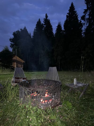 In a grassy field near a forest, a fire pit glows with embers. The pit is surrounded by two wooden chairs and a small folding table. A cabin and trees are visible in the background under a twilight sky. The location is the village of Popovka, Russia.