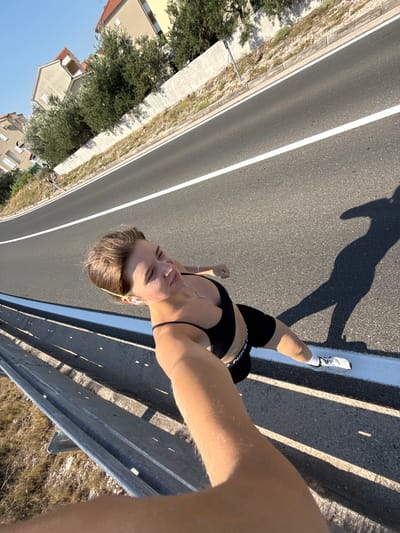 In Grad Šibenik, Croatia, a young woman is captured in a high-angle selfie on a sunny day, outdoors near a road. She is wearing a black sports bra and black shorts, with white athletic shoes and socks. Her hair is light brown and pulled back. She is wearing small white earbuds and a necklace. The woman appears to be mid-stride, possibly running, with her right arm extended to hold the camera. Her shadow is cast on the asphalt road, indicating a strong light source. The road has a white lane marking. To the left of the road, a metal guardrail runs along a slight embankment. Beyond the embankment, a concrete retaining wall is visible, topped with vegetation and trees. In the background, there are residential buildings with reddish-brown roofs and beige facades, nestled amongst green trees, under a clear blue sky. The scene suggests a warm climate and outdoor activity.
