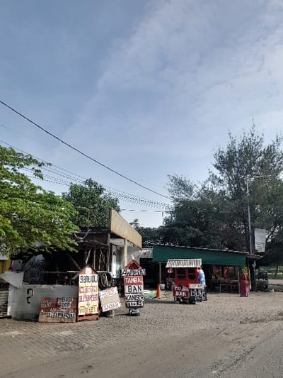 The scene is a roadside shop in Kecamatan Gempol, Indonesia. Several small wooden stalls with signs advertising "ban," "tubes," and "oli garden" are arranged in front of a larger, partially obscured building. There is a single person wearing a red shirt and pants standing near the stall on the right side of the image. The ground is paved with dirt and the scene is illuminated by bright daylight under a slightly cloudy sky. Several power lines run across the sky above the scene. Green foliage and trees can be seen behind and to the sides of the shop.