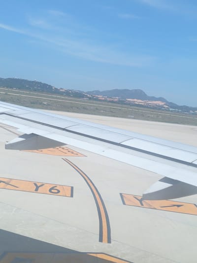 An aerial view shows an airplane wing extending across the foreground. The wing is white and has visible panel lines. On the tarmac below, yellow markings with black lettering indicate taxiway designations. One prominent marking reads "Y6" with an arrow pointing to the right. Another marking shows a single "Y" with an arrow. In the background, a flat landscape with some vegetation and buildings stretches towards a range of low, hazy mountains under a clear blue sky with a few wispy clouds. This scene appears to be on an airport tarmac, possibly near the city of Bắc Cam Ranh, Vietnam, given the context.