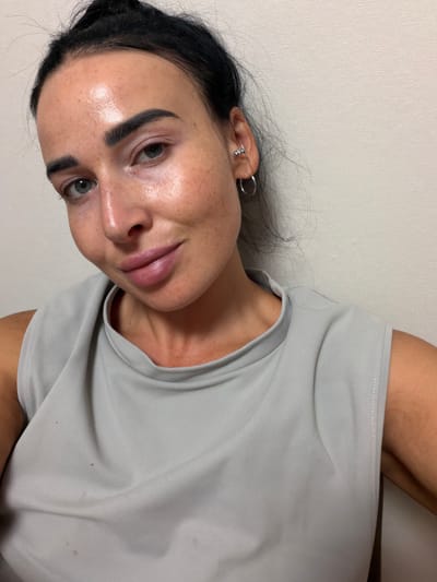 A young woman with dark hair pulled back in a bun, fair skin with freckles, and dark, defined eyebrows is shown in a close-up, selfie-style portrait. She is wearing a light grey, sleeveless top. Her left eye is visible, looking towards the camera, while her right eye is slightly less visible. She has on hoop earrings, one in her left ear with small decorative studs. Her lips are full and appear to be slightly glossy. The background is a textured, light-colored wall. The image appears to be taken indoors. There is no indication of the city of Ladozhskii Trudposelok, Russia.