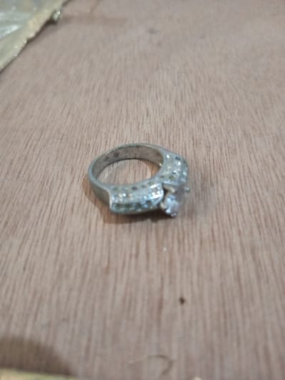 The image displays a silver-colored ring situated on a light brown wooden surface, in the city of Chandragiri, Nepal. The ring features a prominent, clear, faceted central stone secured by four prongs, flanked by sections of the band embellished with multiple smaller, clear, round faceted stones. The metal of the ring appears to be somewhat worn or tarnished. The wooden surface exhibits distinct linear grain patterns and minor surface irregularities. A blurry, light-colored, reflective object is partially visible at the extreme upper-left edge of the frame. No individuals, actions, or interactions are depicted within the scene.