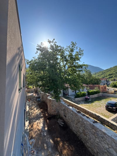 The photograph captures a bright, sunlit outdoor scene in Pelinovo, Montenegro, likely from an elevated perspective. A modern building, possibly under construction or renovation, occupies the left side of the frame, with its light-colored stucco exterior and green-shuttered windows prominently featured. The sun's glare is intense, creating lens flare and casting long shadows across the ground.
In the mid-ground, a stone wall with rough-hewn stones borders a pathway and a yard. Beyond the wall, several buildings are visible, including a small house with a tiled roof and other structures that appear to be in various stages of construction, characterized by exposed brickwork and scaffolding. A large, leafy tree dominates the central part of the image, its branches reaching towards the bright blue sky. In the yard, a pink scooter is parked, and a dark-colored car is visible on the right side. The background reveals a lush, green, mountainous landscape characteristic of the region, with rolling hills covered in trees. The overall impression is one of a rural or semi-rural setting experiencing a clear, sunny day.