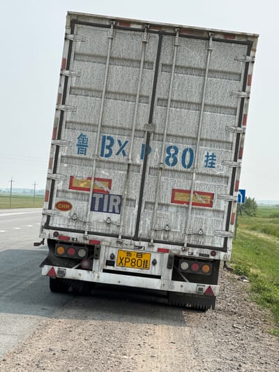 The image shows the rear of a white refrigerated truck trailer parked on the side of a paved road. The trailer is angled, with its rear doors closed. The side of the trailer is marked with blue lettering that reads "BXT" and "80" in a larger font, and some Chinese characters. There are also red and yellow rectangular stickers on the trailer, one with a pictogram of a cigarette with a line through it, indicating no smoking. A yellow license plate with black lettering "XP80 B" is visible on the rear of the trailer, along with "CHN" in a red circular sticker. The truck has red and amber brake lights.
The scene appears to be outdoors, with a dusty, gravelly shoulder next to the road. Beyond the road, there is a grassy field and some trees. The sky is overcast, suggesting a cloudy or hazy day. The time of day is difficult to determine precisely due to the lighting conditions, but it appears to be daytime. The overall impression is one of a parked transport vehicle in a somewhat rural or undeveloped area. There are no people visible in the image. The setting could be a rest stop or a lay-by along a highway in Russia, possibly near Kargat, given the location context. The trailer is somewhat weathered and dirty, showing signs of use.
