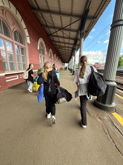 The image, taken in Malaya Vishera, Russia, depicts a group of people waiting on a train platform next to a historic-looking building, under a bright daytime sky. The photograph is rotated 90 degrees clockwise.
In the foreground, a young woman with long, light brown hair tied in a ponytail wears a white t-shirt, dark pants, and a black backpack, standing beside a large, ornate white column with a dark base. Behind her are two younger females, also with long hair. One is dressed in a black tracksuit with a black backpack, carrying a blue bag and pulling a yellow suitcase. The other wears a grey t-shirt, light-colored pants, and a beige backpack, and also has a yellow suitcase. Additional indistinct figures are visible further down the platform.
The train platform features cracked asphalt and a yellow safety line. To the left, a long, red-hued building with classical architectural elements, such as multiple large arched windows with decorative light-colored frames and details, stretches into the distance. Above the building's roof, which appears to be corrugated metal, a bright blue sky with scattered white clouds is visible. Ornate white columns support an overhead structure or canopy above the platform. In the background, railway tracks and a red train or wagon are partially visible. A yellow sign is partially visible in the distance, reading "ПЛАТФОРМА НО1" (PLATFORM NO1). The overall scene suggests travelers waiting for a train in pleasant weather conditions.