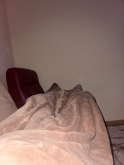 The image depicts an indoor scene in Temse, Belgium, focused from a low perspective. A person is largely obscured by a large, rumpled, brown/taupe plush blanket covering the lower two-thirds of the frame. The blanket conceals the individual's body, with only the top of one foot, clad in a grey sock, visible just above the blanket's edge towards the center-right. To the left, a portion of a light-colored, ribbed fabric furniture piece (possibly a couch arm or cushion) is visible, leading to a dark red or burgundy upholstered recliner or armchair positioned further back. The background consists of a plain, light beige wall. A white electrical outlet or switch plate is visible on the wall in the upper left corner. Scattered small, reflective specks are visible on the wall in the mid-right section. The floor on the far right displays a brown and cream marble-patterned tile. The overall lighting is dim, suggesting an evening or low-light condition.