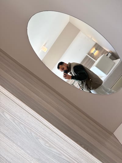 The image depicts a man taking a photograph of himself using a smartphone, reflected in an oval-shaped mirror. The scene is located within a room in Plataniás, Greece.
The man, visible from the chest upwards, has short dark hair and facial stubble. He is wearing a dark green long-sleeved shirt underneath a light tan or olive-colored utility vest with multiple pockets. He holds a black smartphone in his right hand, angled towards himself to capture the reflection.
The mirror's reflection reveals a minimalist room interior, consistent with a hotel room setting. A bed with white bedding and a white upholstered headboard is visible to the right of the man. Adjacent to the bed, a light-colored wall panel features a rectangular, illuminated wall-mounted light fixture. A second similar light is positioned higher on the wall. The room has light-colored walls and a ceiling, from which a square ceiling light fixture is visible.
Outside the mirror's frame, the foreground of the image features a diagonal section of a light-colored wood laminate surface with a distinct horizontal grain, indicative of a wall panel or furniture. The mirror itself is affixed to a light-colored, possibly off-white, wall or ceiling section. The overall composition is angled, with the mirror positioned diagonally across the frame.