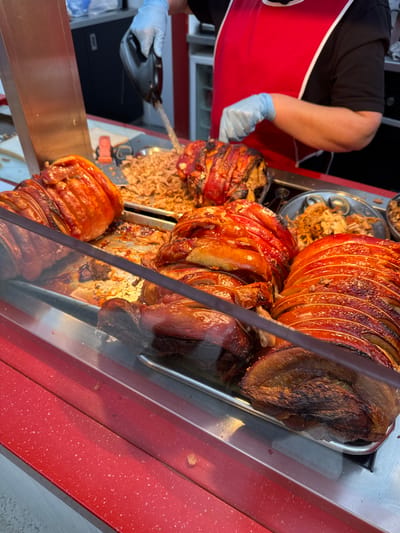 This image captures a food preparation and display counter in Dudley, United Kingdom. In the foreground, a red counter speckled with white flecks runs horizontally, topped by a clear glass barrier. Behind this barrier, multiple large cuts of roasted meat, consistent with pork, are arranged in stainless steel trays. The meat exhibits a reddish-brown, crispy skin and visible fat layers. Several pieces are already pre-sliced into medallions, while one tray contains finely shredded or pulled meat. Liquid drippings and rendered fat are visible within the trays. In the midground, a person wearing a black short-sleeved shirt, a red apron with white trim, and light blue disposable gloves is actively operating a black-handled electric carving or shredding tool. The tool is applied to a large cut of meat, breaking it down into smaller, shredded portions. The background includes light-colored walls and dark cabinetry or storage units.