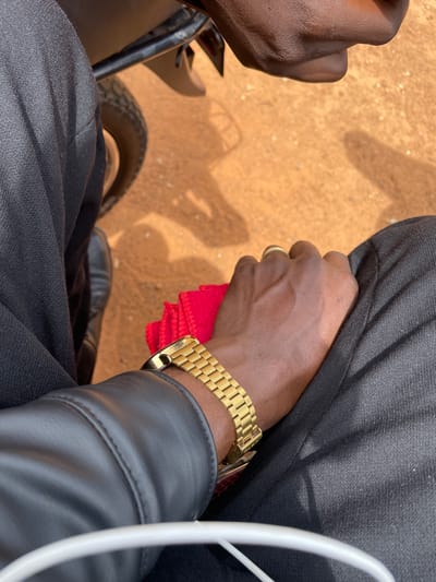 The image shows a close-up of a person's hand and arm, wearing a gold watch. The person is dressed in black clothing, possibly a jacket. A red cloth is visible beneath the hand. The background consists of a sandy, orange-brown surface with shadows cast upon it. This appears to be an outdoor scene, possibly in Kassa, Nigeria, given the sandy terrain and the context of the location.
