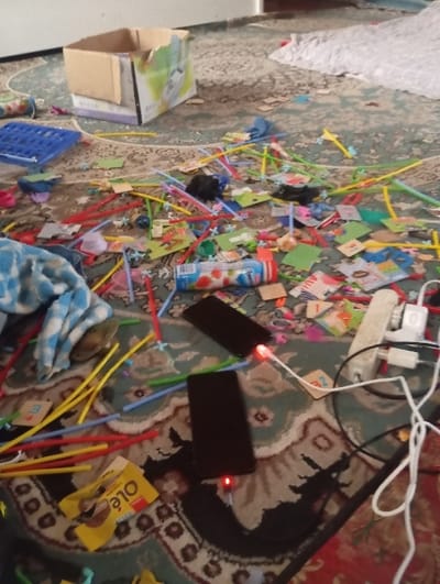 The scene depicts a cluttered indoor floor in Takhtakuprik, Uzbekistan, covered by a patterned rug featuring green, red, and brown tones. Numerous items are scattered across the rug, including a large quantity of colorful plastic straws (red, yellow, green, blue), small plastic connectors, and various small cardboard pieces, some displaying letters or images, consistent with children's educational toys. A blue plastic grid-like toy is also visible. Food-related items present are a plastic bottle with a strawberry-themed label and a yellow "Ole!" wrapper. Two black smartphones are positioned on the rug, both connected to white charging cables, indicated by red lights at their charging ports. These cables lead to a white power strip, which also has a white charger adapter plugged in, along with additional tangled white and black cables. A crumpled blue and white patterned cloth and a small, shiny metallic object are located on the left side of the frame. A partially opened cardboard box is visible towards the rear. No individuals are present in the image.