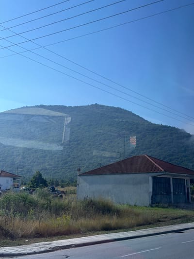 The image captures a rural roadside scene in Kato Nevrokopi, Greece, under a clear blue sky. In the foreground, a paved road with a white stripe runs horizontally across the bottom. Adjacent to the road is a sidewalk composed of light-colored square tiles. Beyond the sidewalk, tall, dry grass and other vegetation line the area.
A low-lying building with a red-tiled roof and a light-colored facade is situated to the right of the frame. This building appears to be a garage or workshop, with large, dark-colored doors spanning its front.
In the middle ground, to the left, a white house with a red-tiled roof is partially visible. The area behind these buildings is a field with scattered dry grass, bushes, and some trees. In the background, a dense, green, tree-covered hillside dominates the landscape, stretching upwards towards the sky. Faint white clouds cling to the lower slopes of the hill. Several power lines stretch across the upper portion of the image, against the backdrop of the bright sky. A rectangular patterned object, possibly a banner or flag, is affixed to the hillside. Reflections are visible on the surface of the image, suggesting it may have been taken through a window.