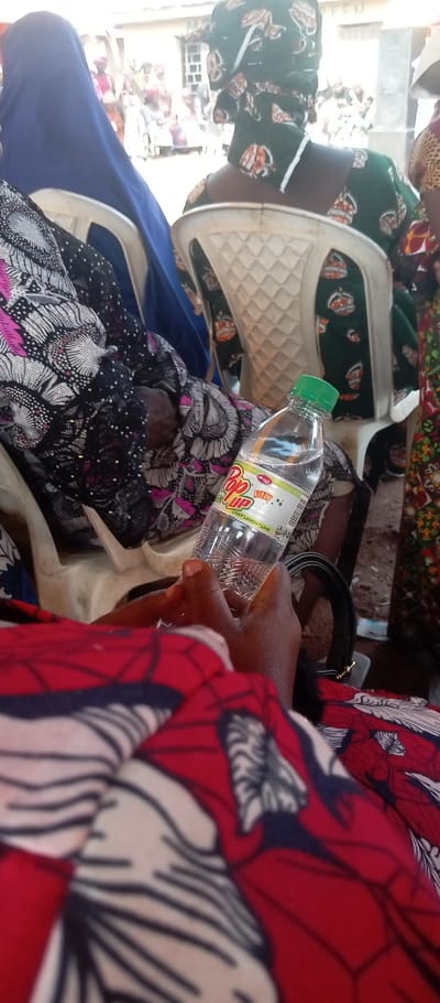 The scene is an outdoor gathering in Kasuwan Magani, Nigeria. The viewpoint is from a seated individual, looking forward and slightly to the right. In the immediate foreground, a person with dark skin holds a clear plastic bottle of "Pop Up Fizzy Light Lemon + Lime" with a green cap. This person wears a red, white, and black patterned fabric. Directly in front, another individual is seated on a white plastic chair, their back to the camera, wearing a dark green patterned dress with orange and white motifs, and a matching headwrap; a white face mask or cloth hangs around their neck. To the left of this individual, another person is partially visible, wearing a blue head covering and a black, white, and pink patterned garment, also seated on a white plastic chair. On the far right, the arm and side of a third person, dressed in multi-colored patterned fabric, are visible. The background reveals a light-colored building with a multi-paned window, and a large dark speaker is positioned to its right. Numerous blurred figures are visible in the distant background, indicating a crowd. The ground appears to be unpaved dirt.