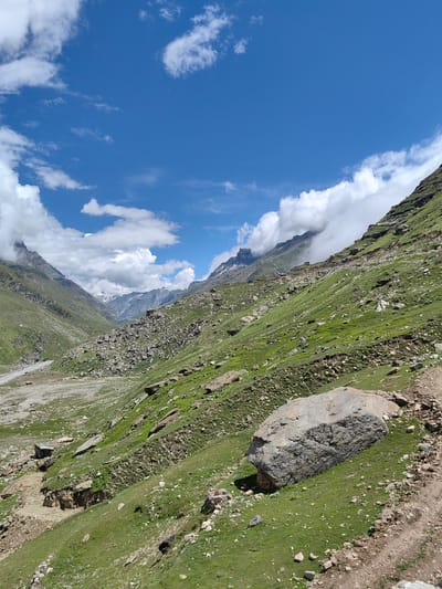 The image captures a panoramic view of a mountainous landscape in Yari-Khoksar, India. The scene is dominated by steep, verdant slopes dotted with numerous rocks and boulders. A winding, unpaved path is visible in the foreground on the right side, cutting across the grassy terrain. In the distance, a valley opens up, revealing a dry riverbed or a gravelly plain, flanked by more mountains. The sky is a vibrant blue, adorned with scattered white clouds, some of which partially obscure the peaks in the background, adding a sense of depth and atmosphere. No people or animals are clearly discernible in the image. The overall impression is one of a rugged, natural environment characterized by its geological formations and expansive sky.