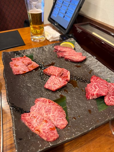 The scene depicts an indoor dining setting within a restaurant in Nagano, Japan. The primary focus is a large, dark, rectangular serving platter with a textured surface, placed centrally on a reddish-brown wooden table. The platter holds multiple distinct groupings of raw, marbled red meat, consistent with various cuts of beef, some resting on small green leaf garnishes. A single yellow lemon wedge accompanies one group of thinner, oblong meat slices on the upper right side of the platter. Several dark brown liquid marks are visible on the platter's surface, distinct from the meat.

To the upper left of the platter, a tall, clear glass filled with an amber liquid and a foamy head, consistent with beer, stands on the table. To the right of the beer glass, a black tablet device, likely an electronic ordering system, is propped upright in a stand, displaying a grid of items with Japanese text on its screen. A small, folded white cloth or paper towel is situated between the beer glass and the tablet. Further to the right, a dark, rectangular wooden box with a clear top, possibly a chopstick holder, reveals a light-colored object within. A metallic utensil, potentially a pair of tongs or chopsticks, is partially visible on the far left edge of the table. The background shows blurred details of a dark patterned chair, indicating a seating area. No individuals are visible in the frame.