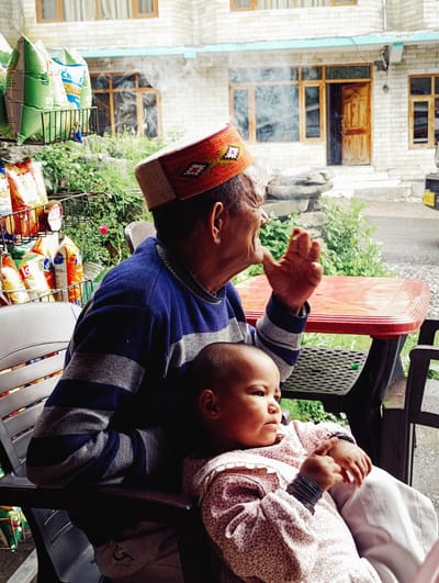 In Kothi, India, an elderly man wearing a traditional cap with red and green embroidery is seated on a chair. He is smoking and exhaling a plume of smoke. Next to him, a young child is also seated. The child is wearing a light-colored garment with a repeating pattern and bracelets on their wrists. Both individuals are positioned near a red plastic table. Behind them, a rack displays various snack bags. The background shows a building with brick walls and wooden windows, along with greenery.