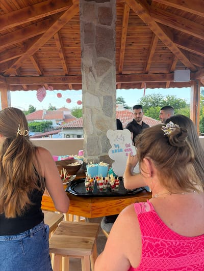 The image depicts an outdoor gathering, likely a baby shower, held on a wooden patio or gazebo with a stone pillar supporting a wooden roof. The location is identified as the city of Troyanovo, Bulgaria. In the foreground, a woman with long brown hair tied back with a gold clip is seen from behind, wearing a black tank top and blue denim shorts. She is seated at a wooden table laden with food, including popcorn in blue cups, skewers of appetizers, and bowls of snacks.
Further into the scene, other individuals are present. Two men, one wearing a black polo shirt and the other a black t-shirt, are standing near a decorative "What will baby BE?" board shaped like a bear. This board has sections for "Boy" and "Girl" with small blue and pink cutouts, suggesting a gender reveal element. A woman with her hair in a bun, adorned with a floral accessory, is also interacting with the board, pointing towards it. Another woman with long dark hair is partially visible in the background, standing next to the men.
The backdrop reveals a residential area with tiled roofs and greenery, under a bright sky with some clouds. Pink paper fans and circular decorations are suspended from the wooden structure, adding to the festive atmosphere. The overall scene suggests a casual, celebratory event taking place during daylight hours.