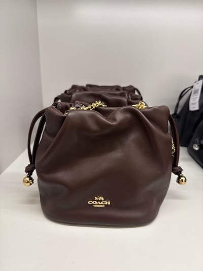 The image shows a brown Coach drawstring bag with the Coach logo on its front. The bag is sitting on a white surface, likely a shelf or display case. The picture was probably taken indoors, and there is no indication that it was taken in Sunrise, United States of America.