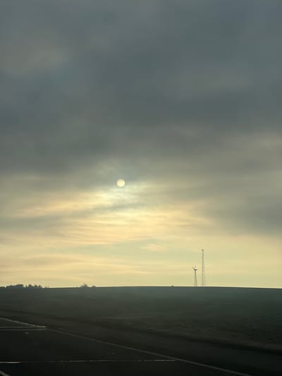 The image shows a landscape in Вольнаўскі сельскі Савет, Belarus, under an overcast sky with the sun obscured by clouds. A road runs along the right side of the frame. Adjacent to the road is a dark field with a tree line visible. In the distance, there is a cell tower and a wind turbine. The sky transitions from a dark grey on the left to a lighter, yellowish hue on the right where the sun is located.