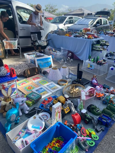 This is a medium shot, taken at an outdoor market in San Luis De Sabinillas, Spain. The market appears to be a flea market or car boot sale, with items laid out on tarps and tables, some of which are set up next to parked cars. The ground is paved with asphalt and gravel.

In the foreground, a variety of toys and trinkets are displayed on a blue and grey tarp. This includes several stuffed animals, a blue toy robot, toy cars, building blocks, a toy keyboard, and what appears to be a roulette wheel. A blue plastic bin is filled with colorful building blocks. Next to it, a white bin contains smaller toys and packaging.

Further back, a table is covered with a white tablecloth, displaying more toys, including playmobil figures, toy vehicles, and what looks like a toy house. Behind this, more tables and tarps are visible, laden with various items for sale, suggesting a wide range of goods are on offer at the market.

Two women are present. One woman, wearing a white tank top and a straw hat, is standing by the open door of a white van, sorting through a bag of items. She is wearing dark trousers and sandals. The other woman, seated in the van, is wearing a sleeveless olive-colored top and dark trousers. She is reaching into a clear plastic bin.

The overall atmosphere suggests a sunny day, with a few tents and parked cars in the background, indicating a typical market scene. The backdrop includes some trees and buildings, and in the distance, mountains can be seen under a bright sky.