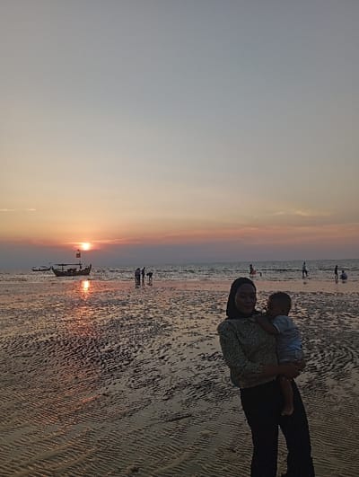 The image depicts a sunset scene on a beach in Tritunggal, Indonesia. In the foreground, a female subject, wearing a dark hijab, a light-colored patterned long-sleeved shirt, and dark trousers, stands facing slightly left of the camera. She holds an infant in her left arm; the infant, dressed in light-colored clothing, faces left, away from the camera. The female subject has a faint smile. The ground is a wet, sandy beach with pronounced ripple patterns, reflecting the sunset's orange light. In the midground, several individuals are scattered across the beach and in the shallow water, primarily appearing as dark silhouettes against the brighter background. Two boats are visible on the water, one being a larger traditional vessel with a mast positioned to the left of the image center. The sun, a distinct orange-red orb, is partially visible on the horizon in the left-center, casting a bright reflection across the water and wet sand. The sky transitions from an orange-red hue near the horizon to a pale blue-grey overhead, with some faint cloud formations near the horizon.