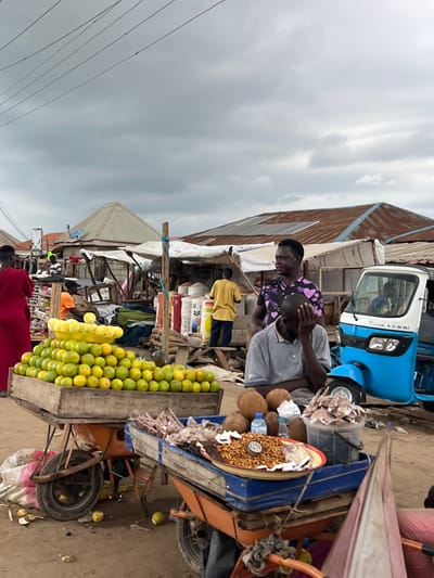 A street market scene unfolds in Oko Amakom, Nigeria, under an overcast sky. In the foreground, a wooden cart overflows with a vibrant display of oranges, neatly stacked in a yellow plastic basin. To its right, another cart holds various goods, including a platter of nuts, coconuts, and a bucket of what appears to be dried fish. A blue and white motorized tricycle, commonly known as an "okada" or "kekeno," is parked nearby.
Several individuals populate the scene. A man in a grey polo shirt sits with his head in his hand, seemingly dejected, next to the tricycle. Another man with a purple shirt adorned with hearts stands behind him, looking towards the left. To the far left, a person in a bright red garment stands facing away from the camera. In the mid-ground, a person in a yellow shirt is visible standing near a stall. The market environment is characterized by makeshift stalls with corrugated metal roofs and tarpaulin coverings, typical of informal markets in the region. Wires are visible strung overhead. The ground is dusty and uneven, with some scattered debris. The overall atmosphere suggests a bustling, albeit somewhat subdued, market day.