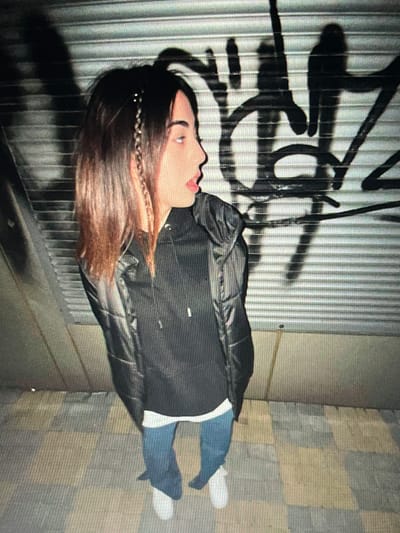 A young woman with brown hair styled in a braid with beads stands in front of a corrugated metal wall covered in black graffiti. She is wearing a black puffer jacket over a black hoodie and blue jeans. The ground is paved with square tiles in shades of grey, yellow, and brown. The lighting suggests it is nighttime or a dimly lit indoor space.